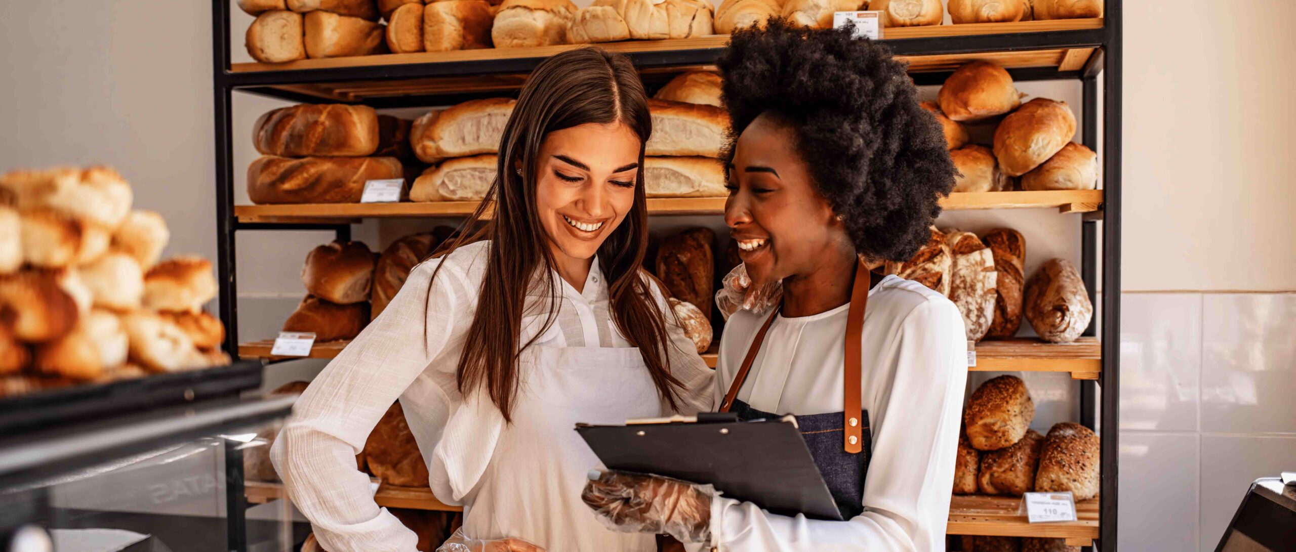 Na imagem duas mulheres sorridentes, com prateleiras com pães ao fundo. Uma está segurando uma prancheta, enquanto a outra olha para a prancheta.